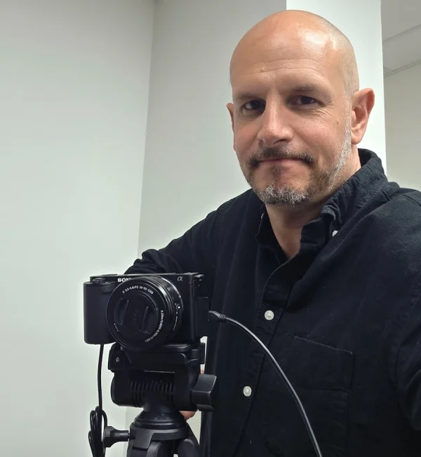 Headshot of Neal Veglio standing next to a camera on a tripod
